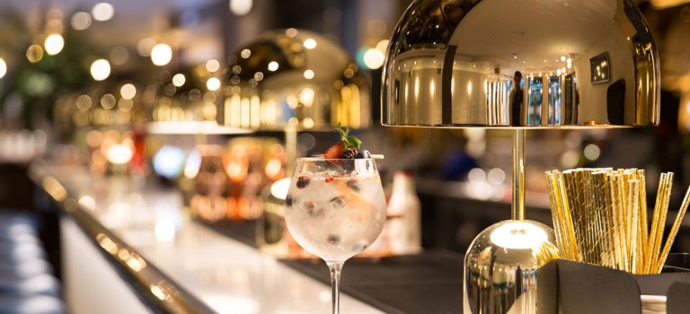 Gekonnt kontern: Energiepreise Getränk mit Beeren auf Tresen der Hotellerie neben Strohhalmen aus Edelstahl und goldenen Tischlampen